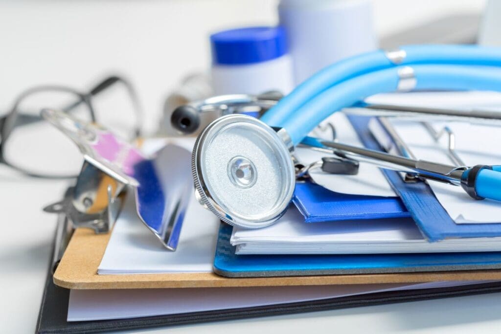 A blue stethoscope, a clipboard, documents, eyeglasses, and medical containers are arranged on a desk, suggesting a healthcare or medical setting.