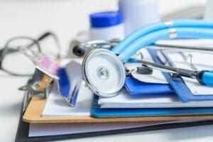 A blue stethoscope, a clipboard, documents, eyeglasses, and medical containers are arranged on a desk, suggesting a healthcare or medical setting.