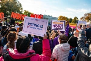 A crowd gathers outdoors holding signs advocating for voting rights, including one that reads "Voting is People Power," near the White House.
