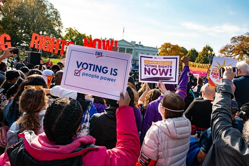 A crowd gathers outdoors holding signs advocating for voting rights, including one that reads "Voting is People Power," near the White House.